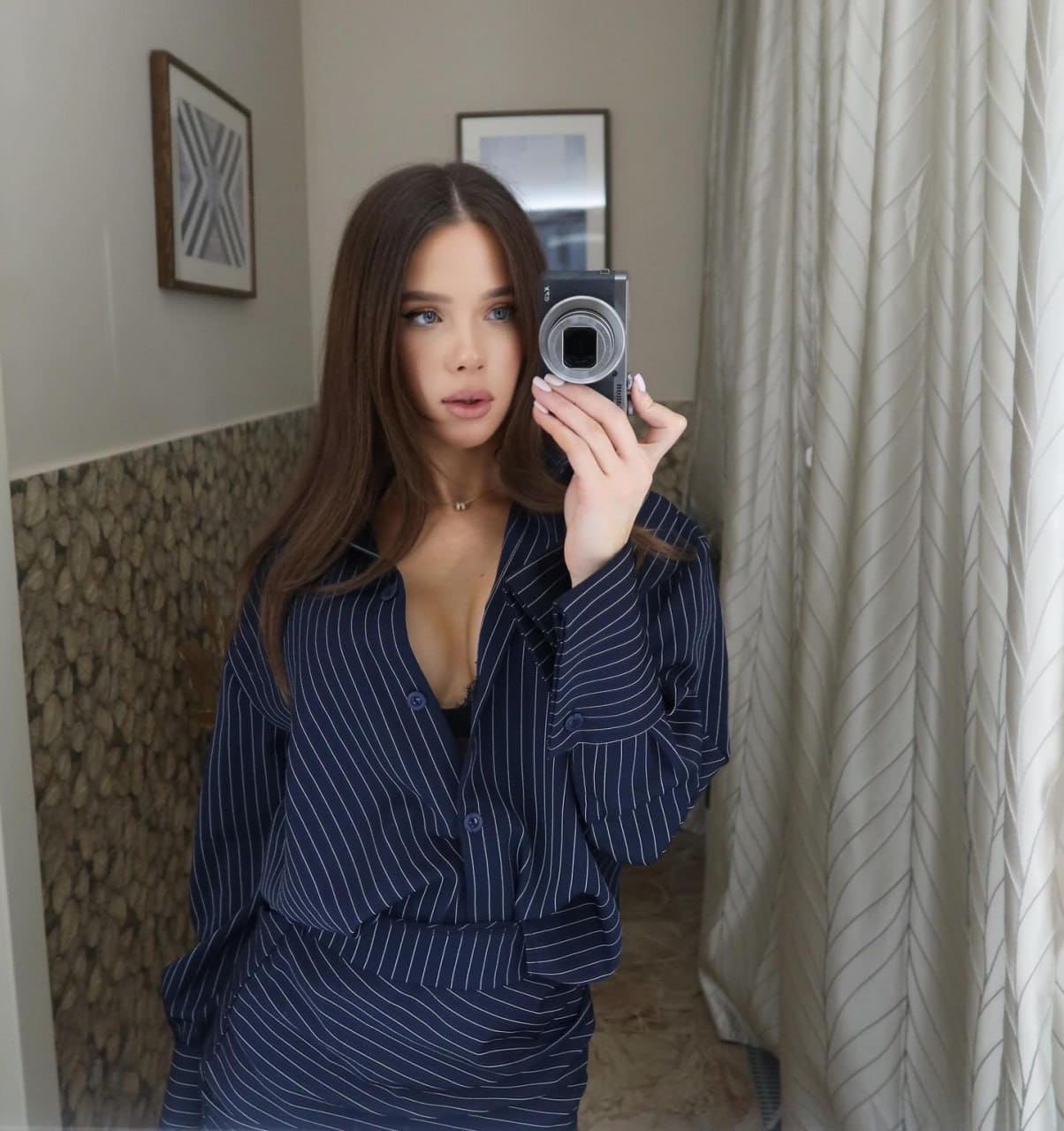 A woman takes a mirror selfie in a room with framed pictures, wearing an open dark blue pinstriped shirt over a black top.