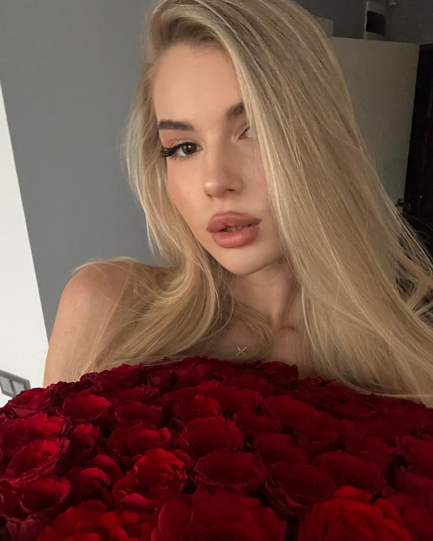 A woman holds a large bouquet of red roses covering her lower body, with bare shoulders visible against a plain indoor background.