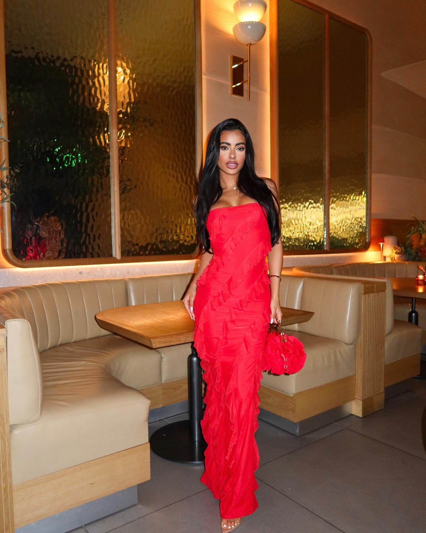 Christie stands in a restaurant booth wearing a strapless, floor-length red ruffled gown and holding a matching red handbag.
