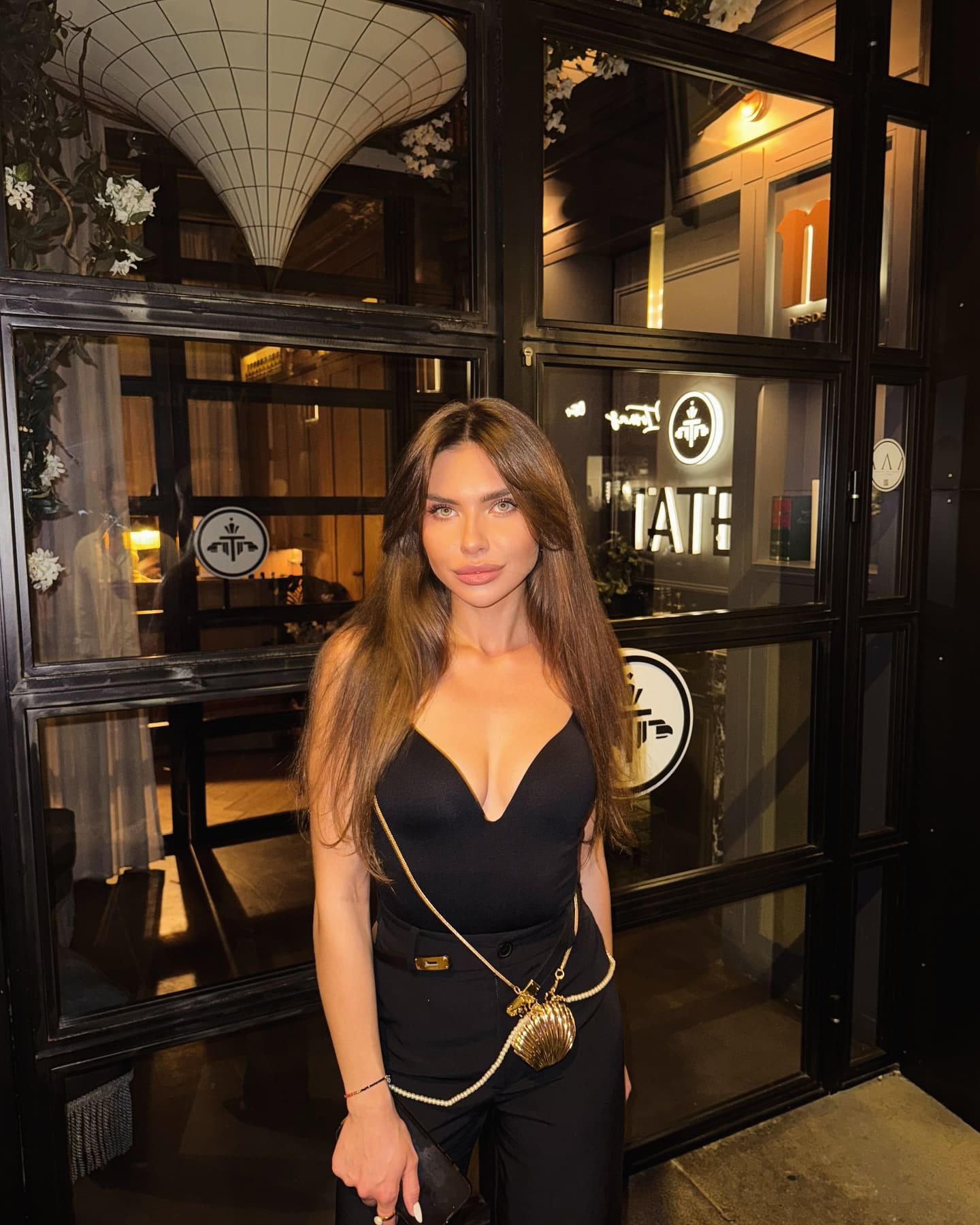 Safira stands at night in front of a glass storefront, wearing a black V-neck top, trousers, and a gold crossbody bag.