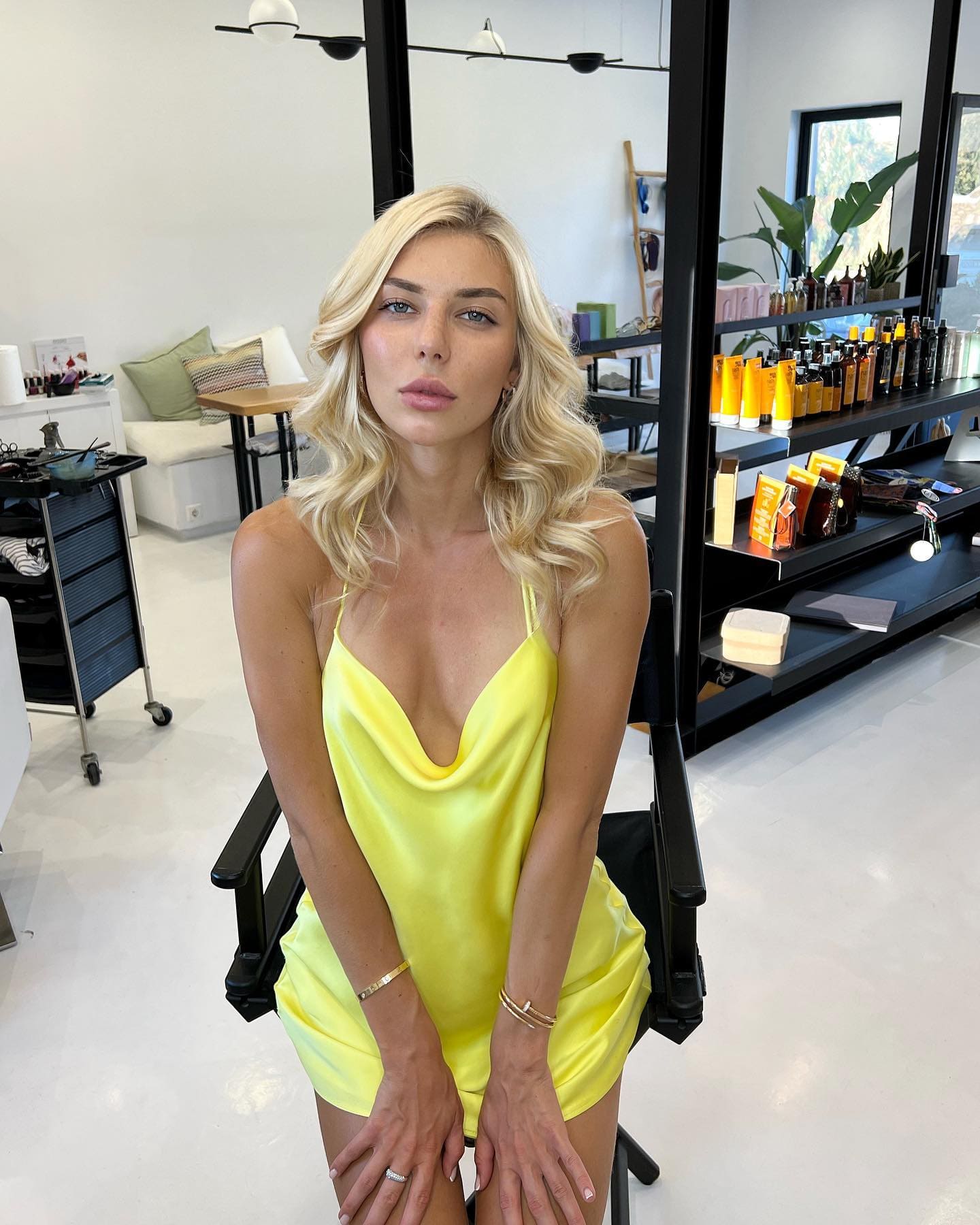 Inga sits in a black chair wearing a short yellow satin slip dress, with shelves of hair products and a mirror visible in the bright room behind her.