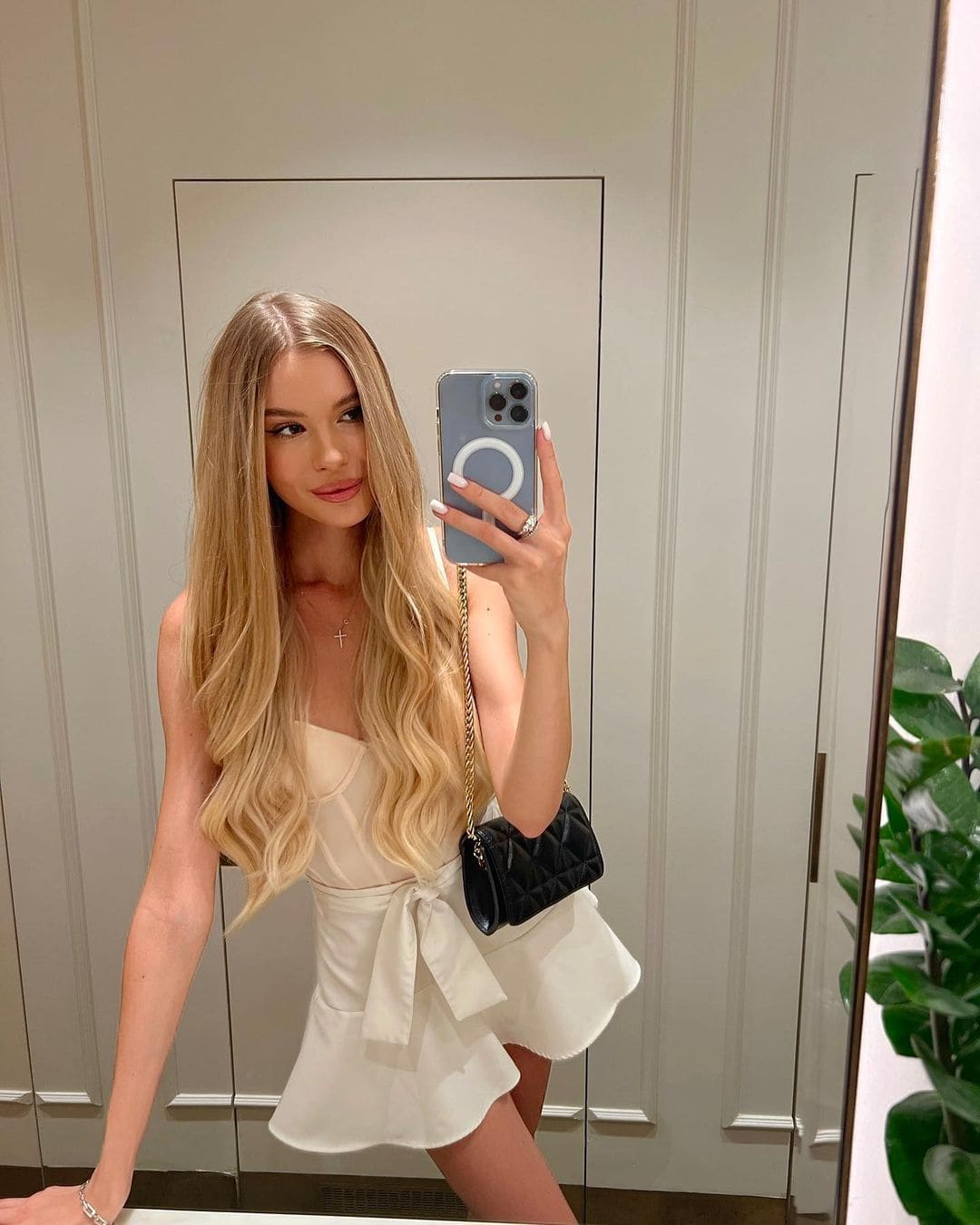 Rita takes a mirror selfie in a white sleeveless dress and black shoulder bag, standing before a paneled wall with a potted plant.