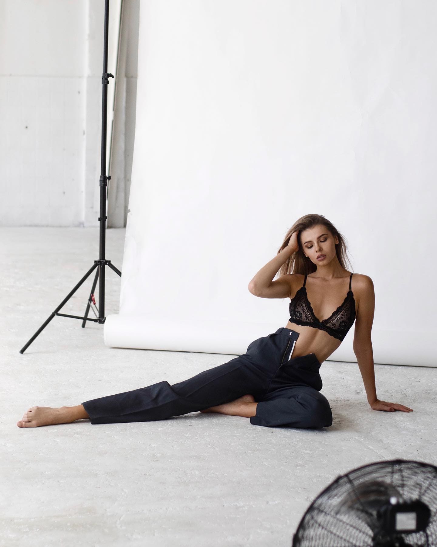 A woman with brown hair sits on a concrete floor in a black lace bra and trousers, hand in hair, with a lighting stand visible behind her.