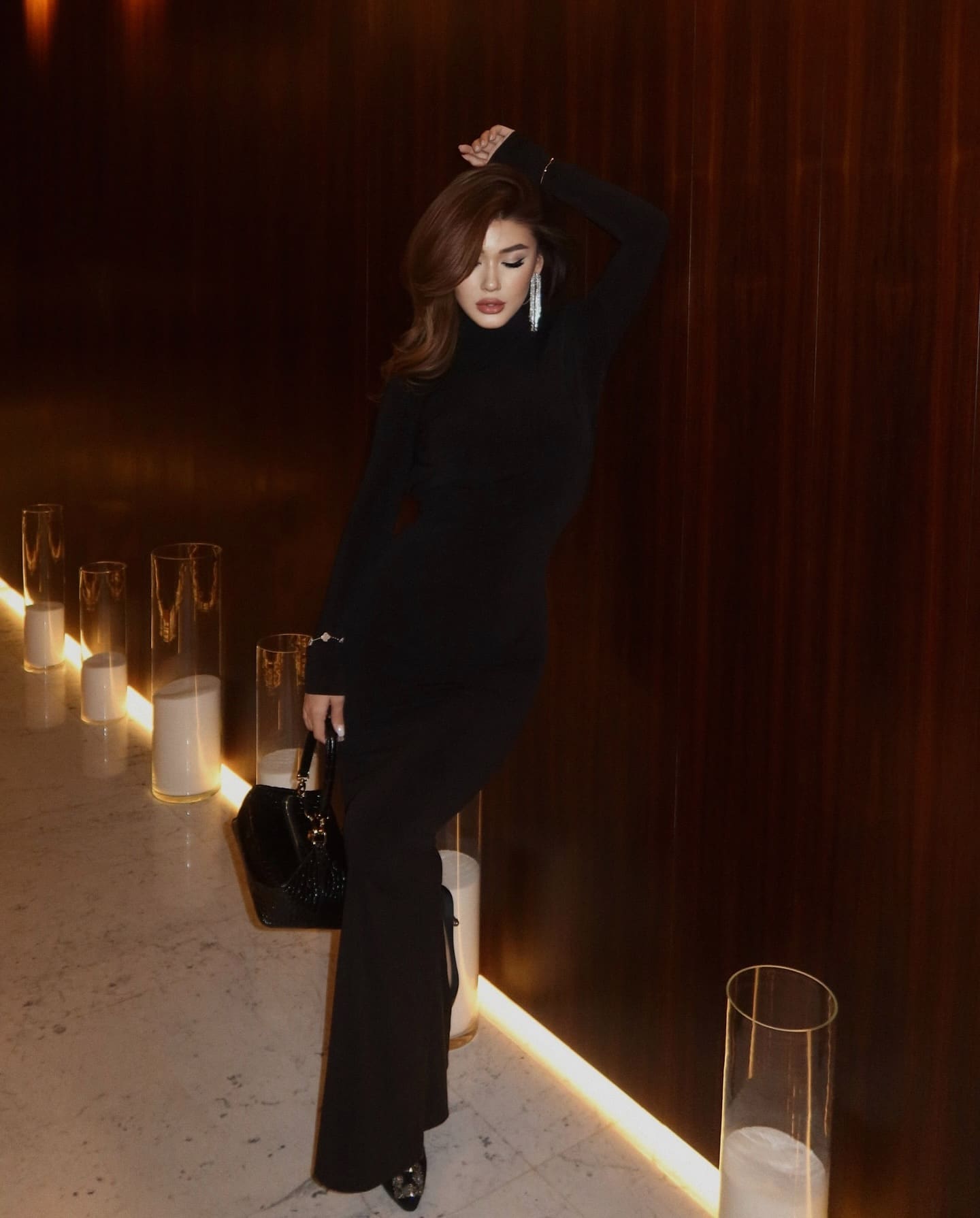A woman in a black long-sleeved high-neck flared dress poses in a dimly lit room with a dark wood wall, holding a black handbag with one hand raised.