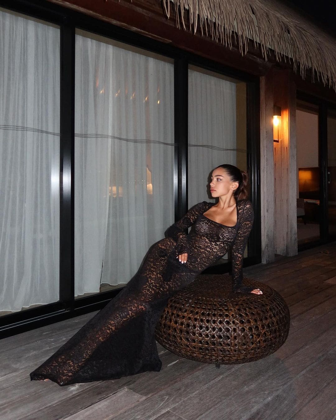 A woman sits on a woven ottoman in a black lace gown, legs extended, near curtained windows and a thatched roof.