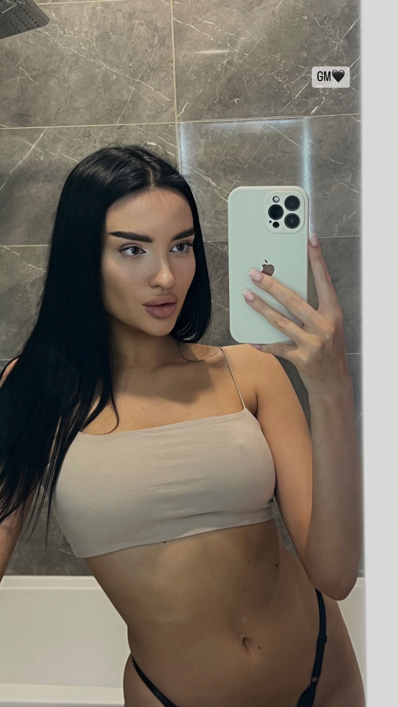 Mirror selfie in a tiled bathroom showing a woman in a beige bandeau top and black underwear, holding a light-colored smartphone.