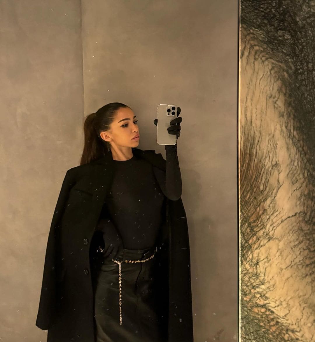 A woman in a black coat, gloves, and chain belt takes a mirror selfie with a smartphone against a plain wall.