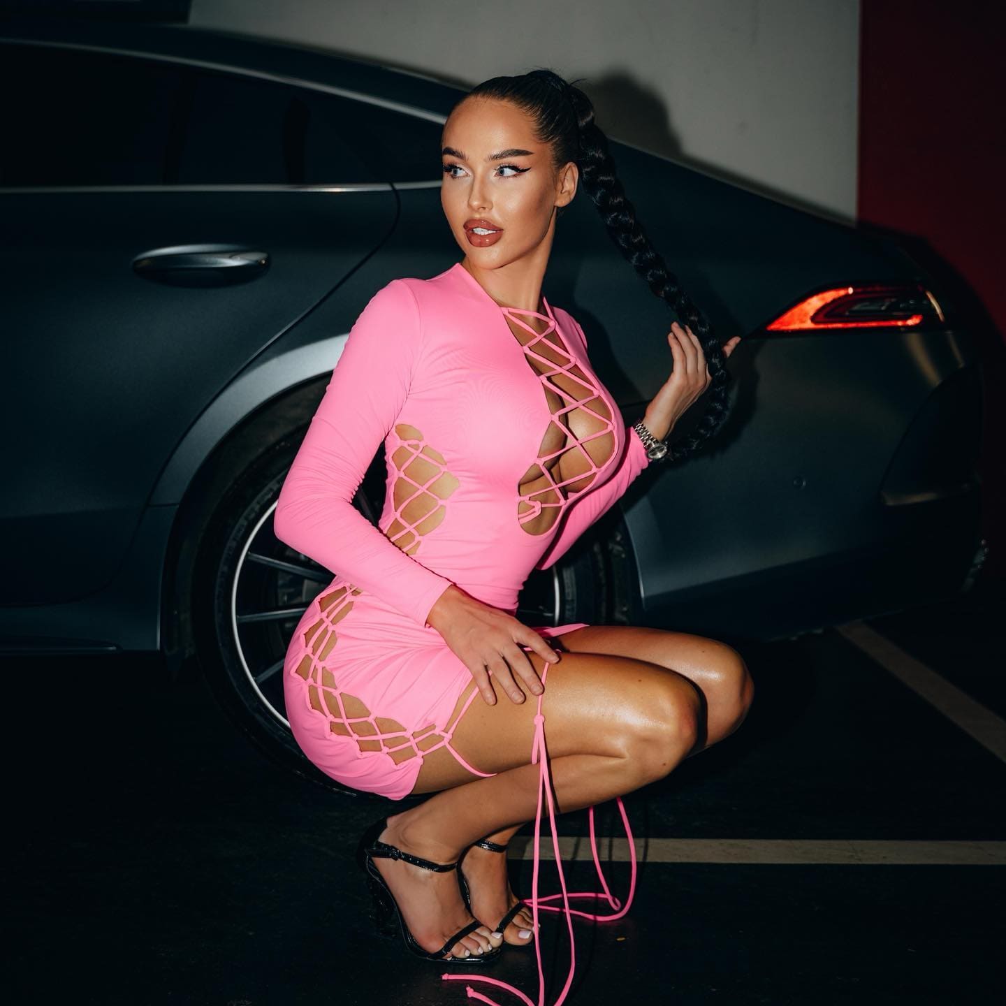 Crouching before a dark car, she wears a pink mini dress with cutouts and strappy heels.