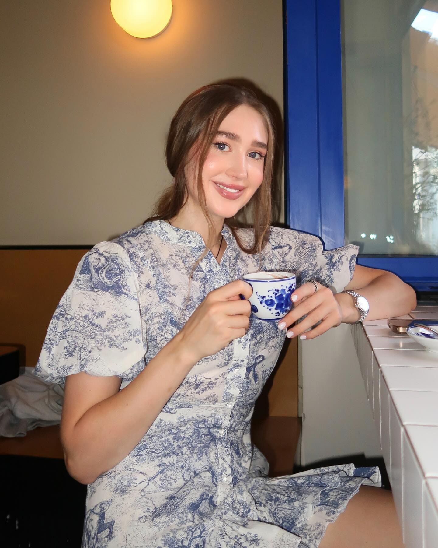Aeneas sits at a white tiled counter in a blue floral dress, holding a patterned teacup, with a blue-framed window behind her.