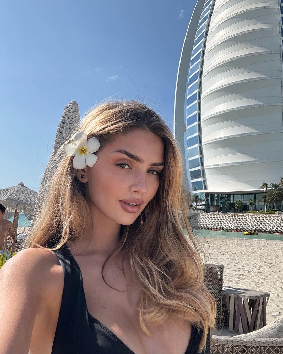 Olga poses on a sandy beach in a black sleeveless top, with a white flower behind her ear and a curved building in the background.