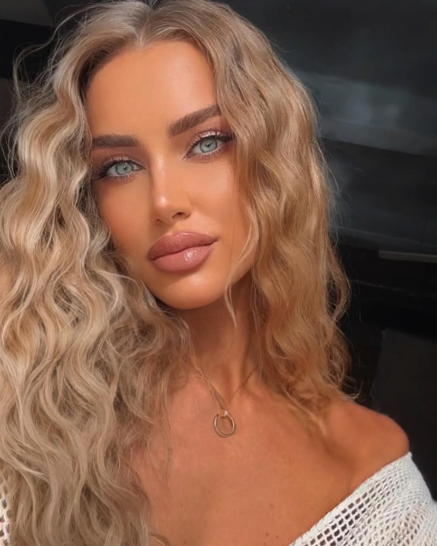 Milena with long wavy blonde hair wears a white off-the-shoulder knit top and circular pendant necklace against a dark blurred background.