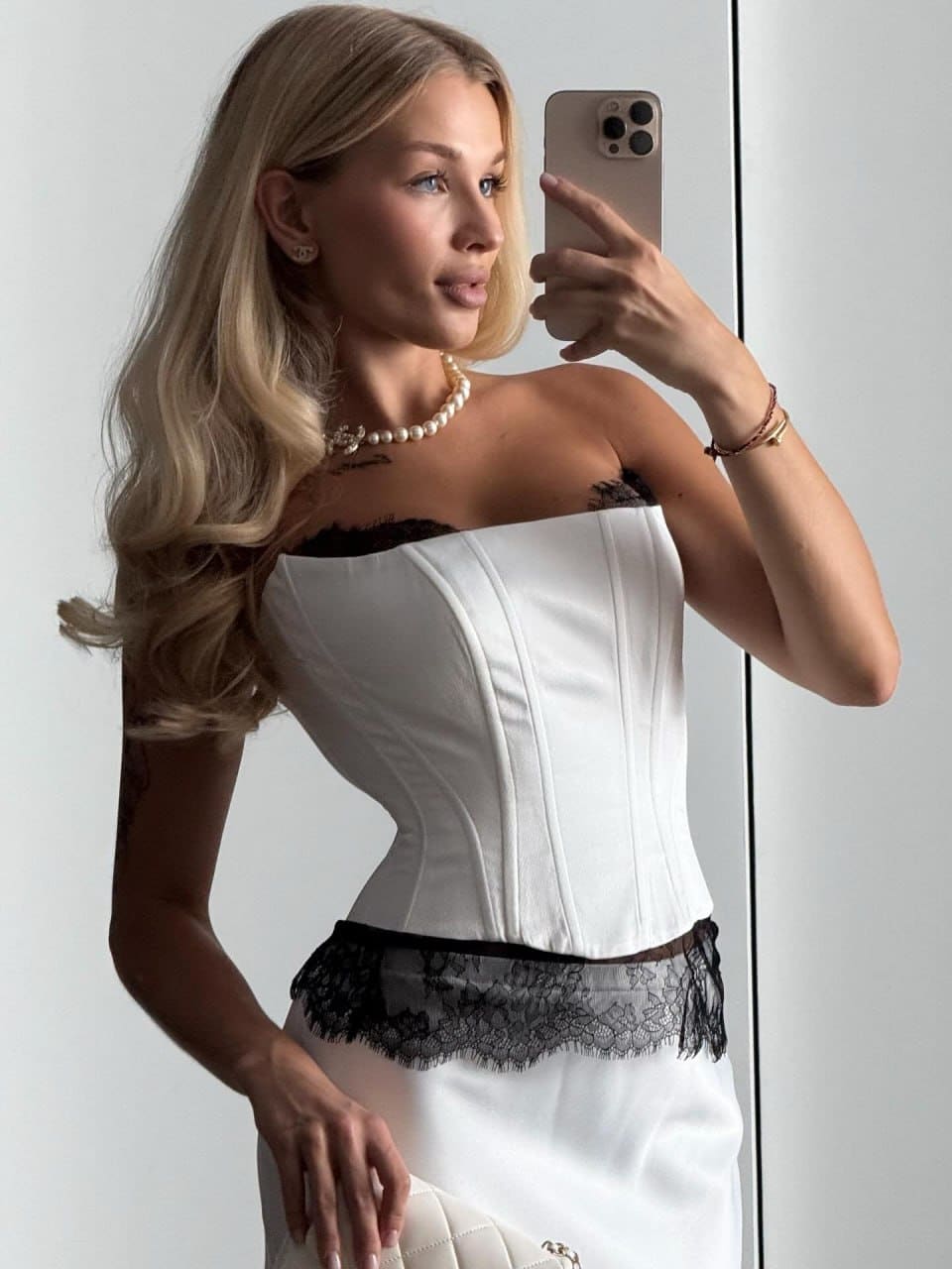 A mirror selfie shows a woman in a white corset top with black lace trim and white skirt, wearing a pearl necklace against a light wall.