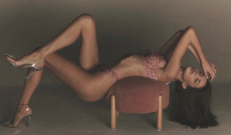 A woman lies on her side on a stool in pink patterned lingerie and clear heels, arm raised over her face against a gray background.