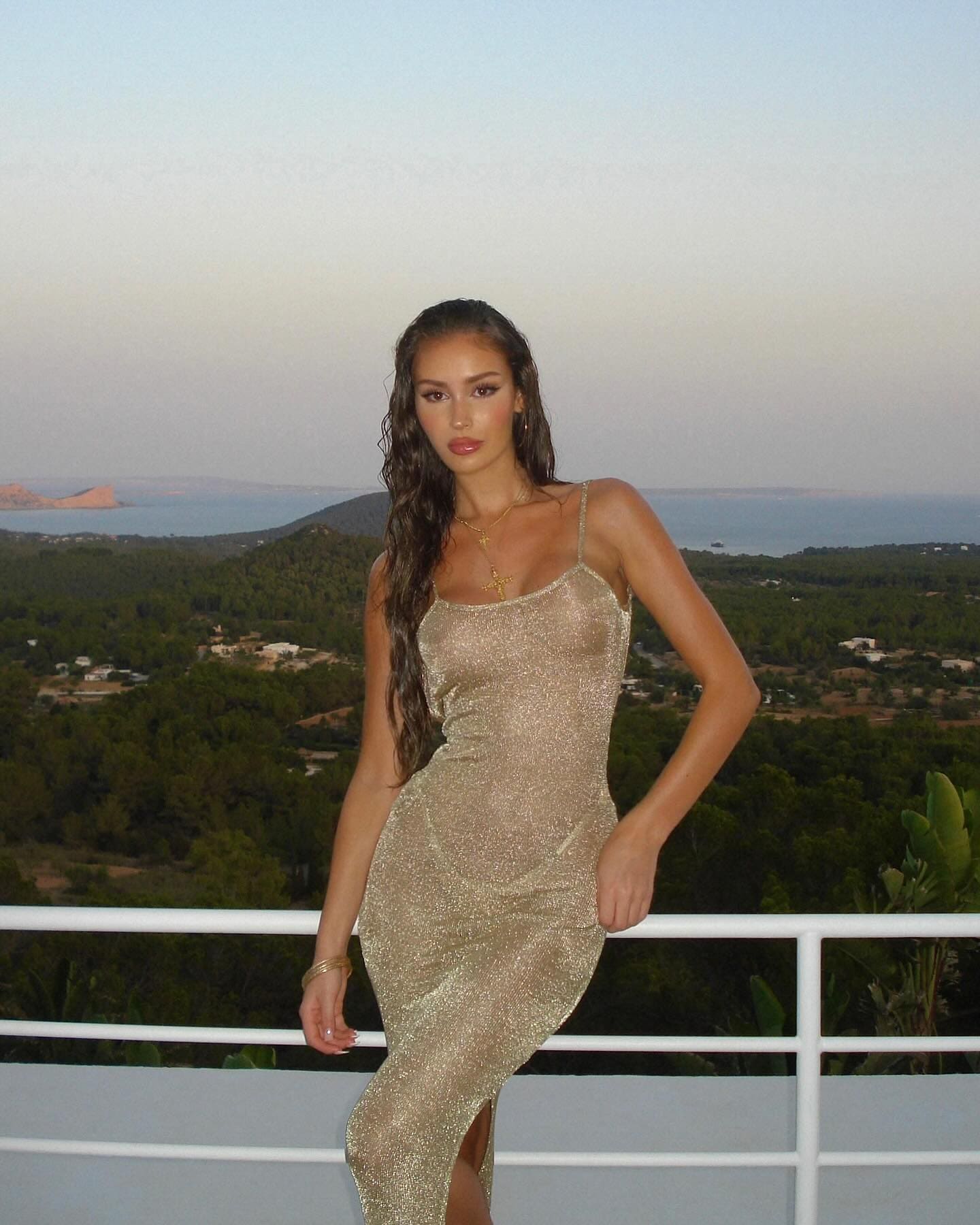 On a balcony with a white railing, a woman in a shimmering gold slip dress poses against a backdrop of green hills and distant water.