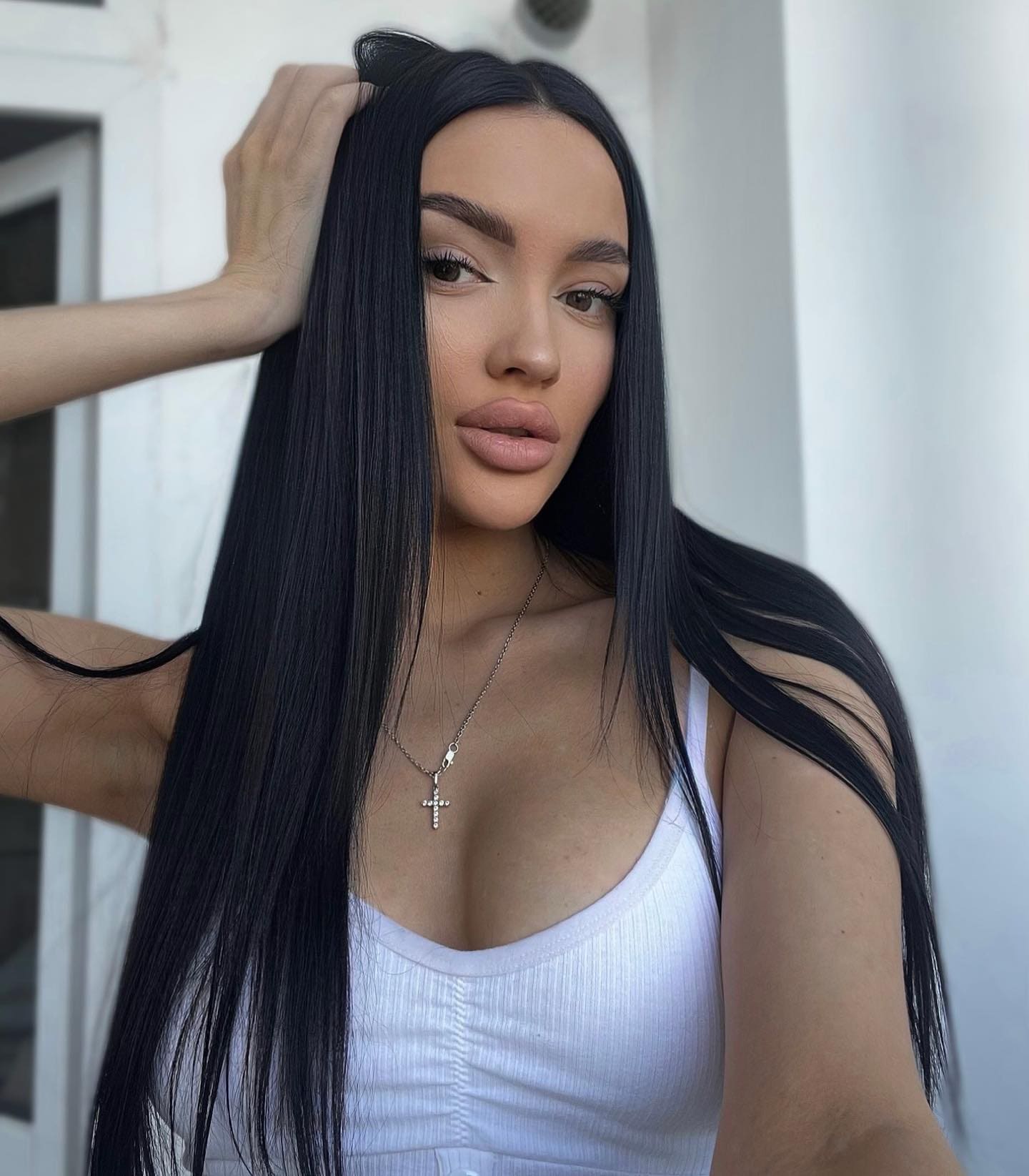 Iza stands outdoors against a white wall, wearing a white ribbed tank top and cross necklace, holding her long black hair up with one hand.