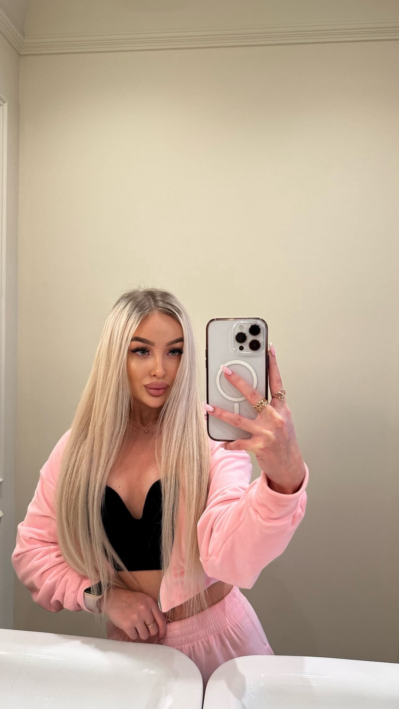 In a bathroom mirror selfie, she wears a pink cropped jacket over a black bralette and pink sweatpants, holding a white-cased phone.