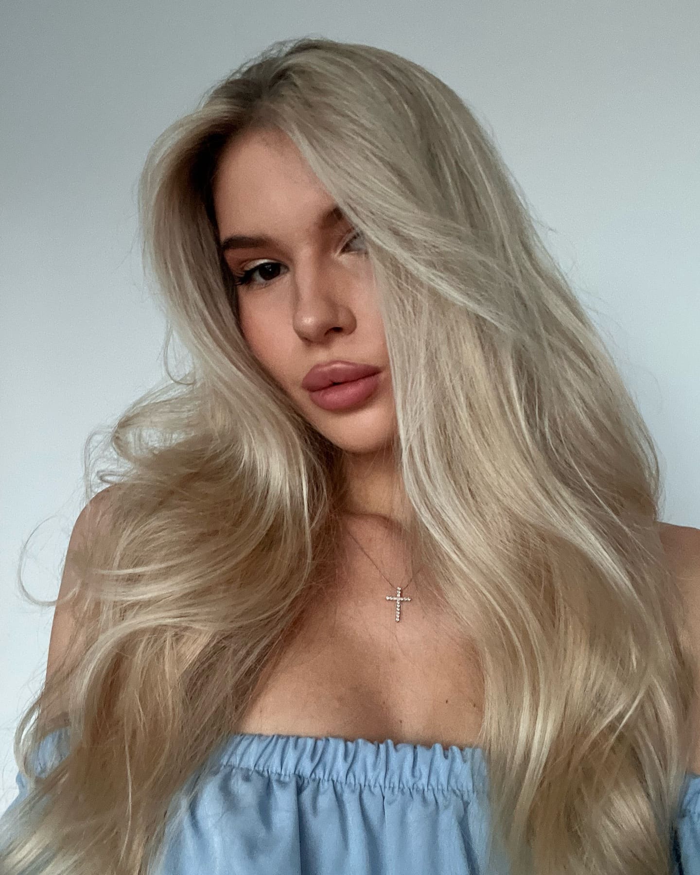 A woman with platinum blonde hair wears a light blue off-the-shoulder top and cross necklace, posing against a plain light background.