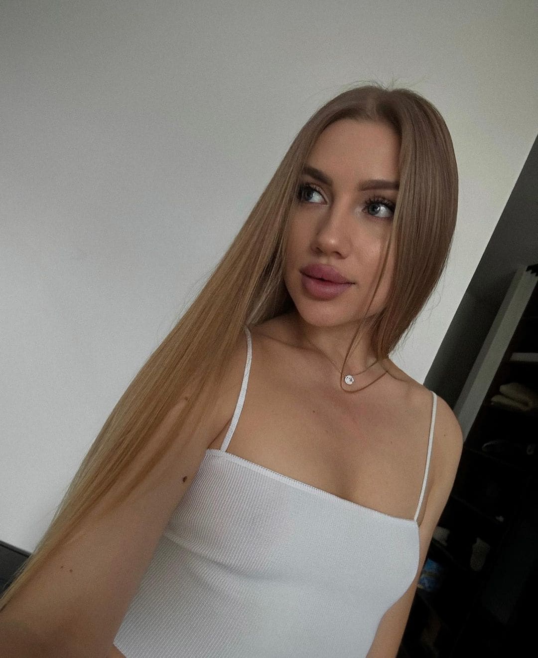 Posing indoors in a white ribbed spaghetti-strap top and necklace, looking to the side against a plain wall.