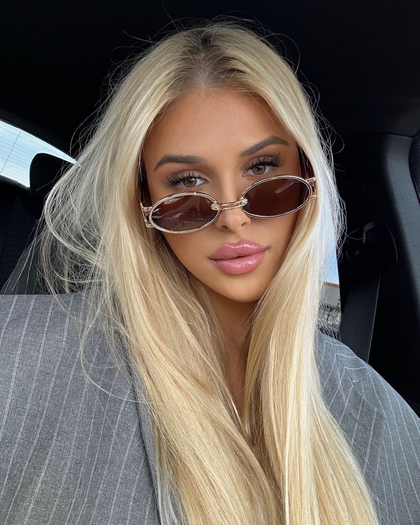A blonde woman sits in a car back seat, wearing gold-framed oval sunglasses and a grey pinstriped blazer.
