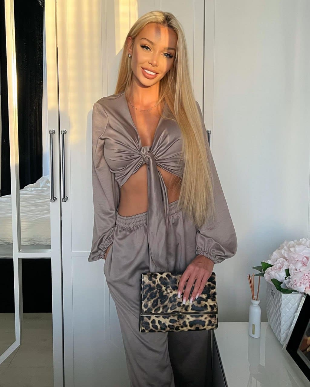 Naomi stands indoors in a taupe satin two-piece outfit with a knotted top and trousers, holding a leopard-print clutch in front of white wardrobes.