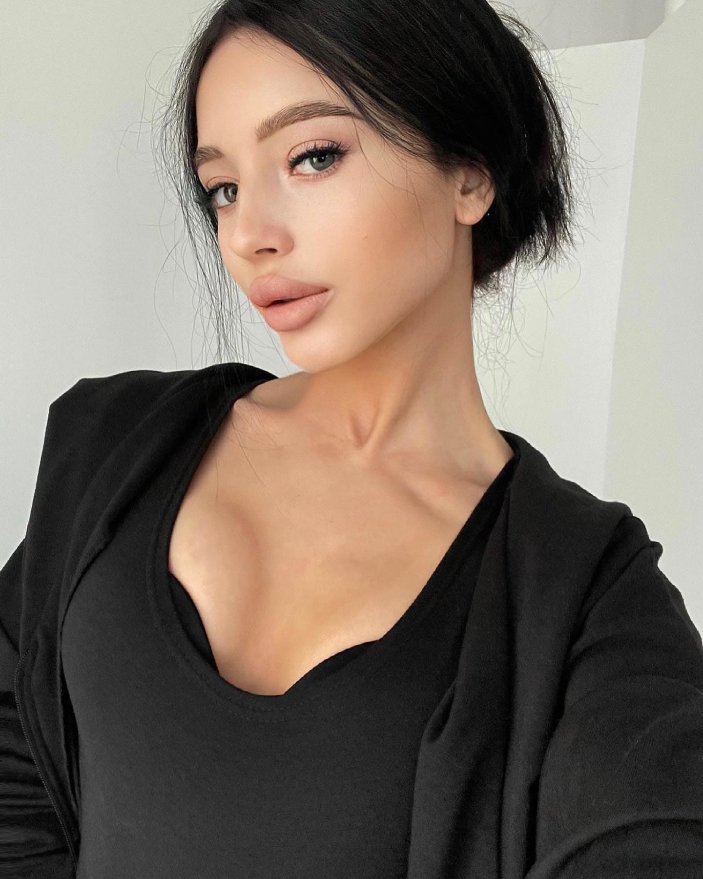 Maryana with dark hair pulled back wears a black scoop-neck top and cardigan, posing against a plain light wall.