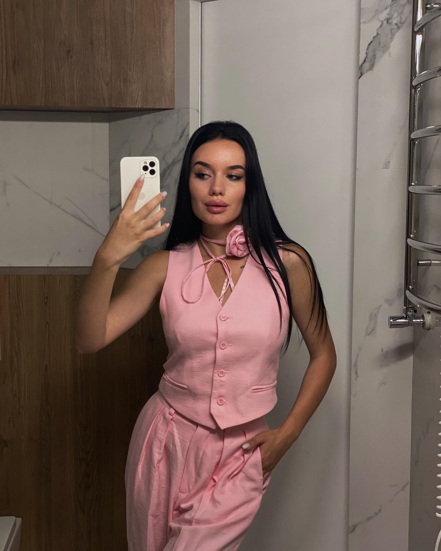 Julia takes a mirror selfie in a marble bathroom, wearing a pink floral button-up top and trousers, holding a white phone.