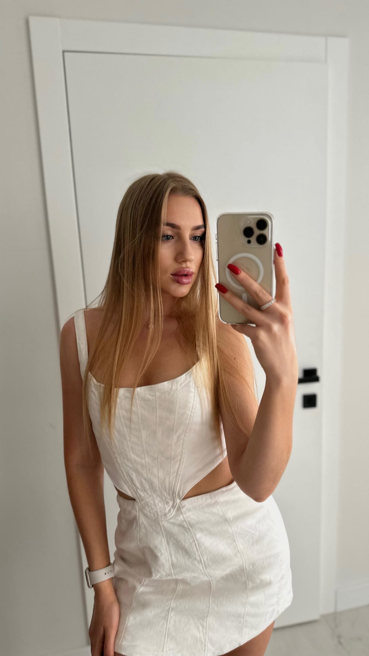 Mirror selfie in a white sleeveless top and short skirt, standing before a white door.