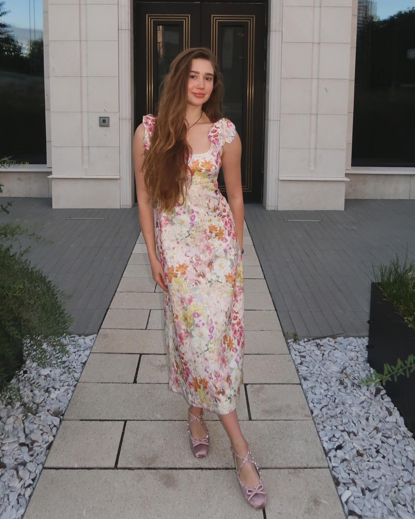 A woman with long wavy brown hair stands on a stone walkway in a floral midi dress and pink strappy flats.