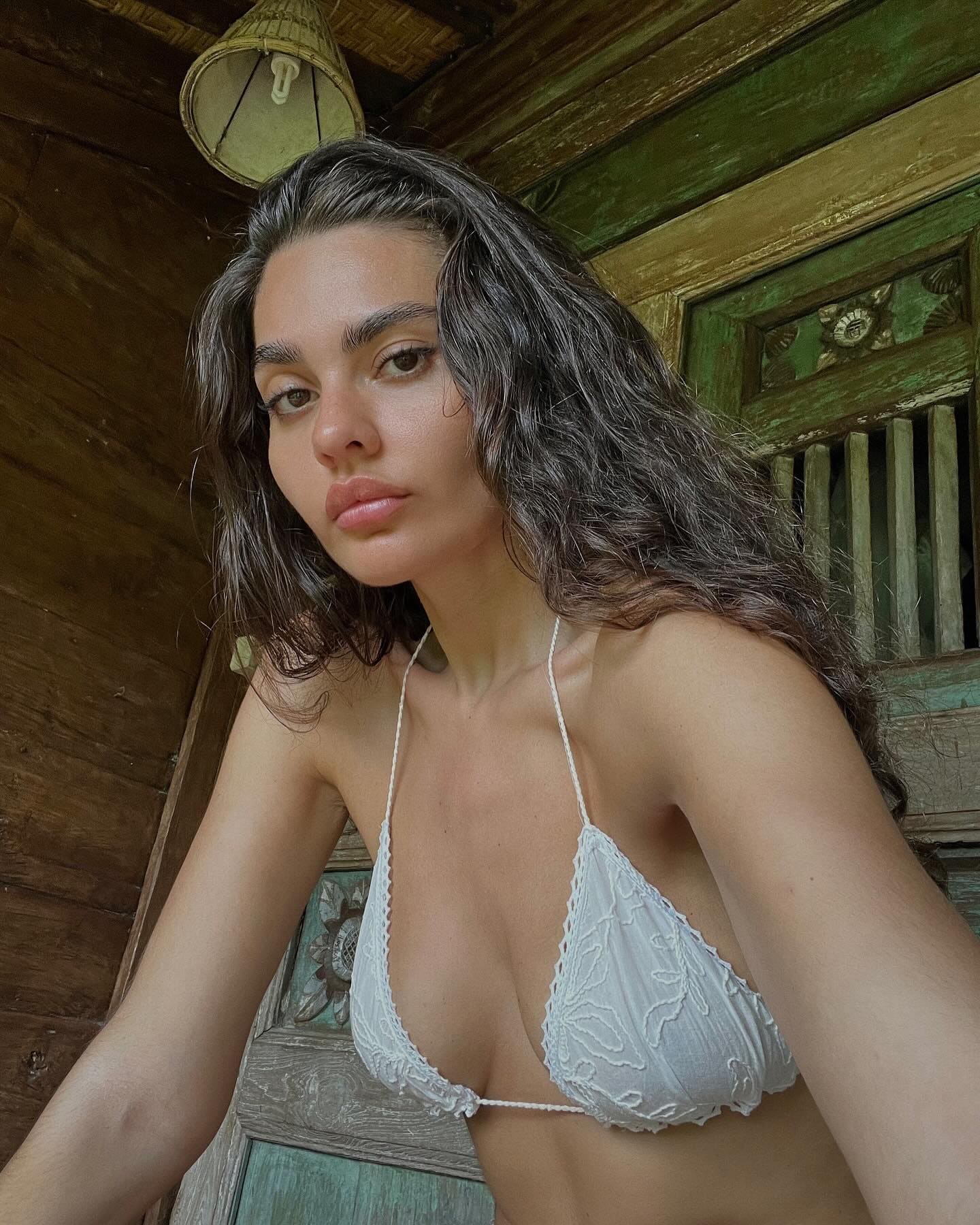 A woman in a white lace bralette poses before a weathered wooden wall and a green-painted window frame.