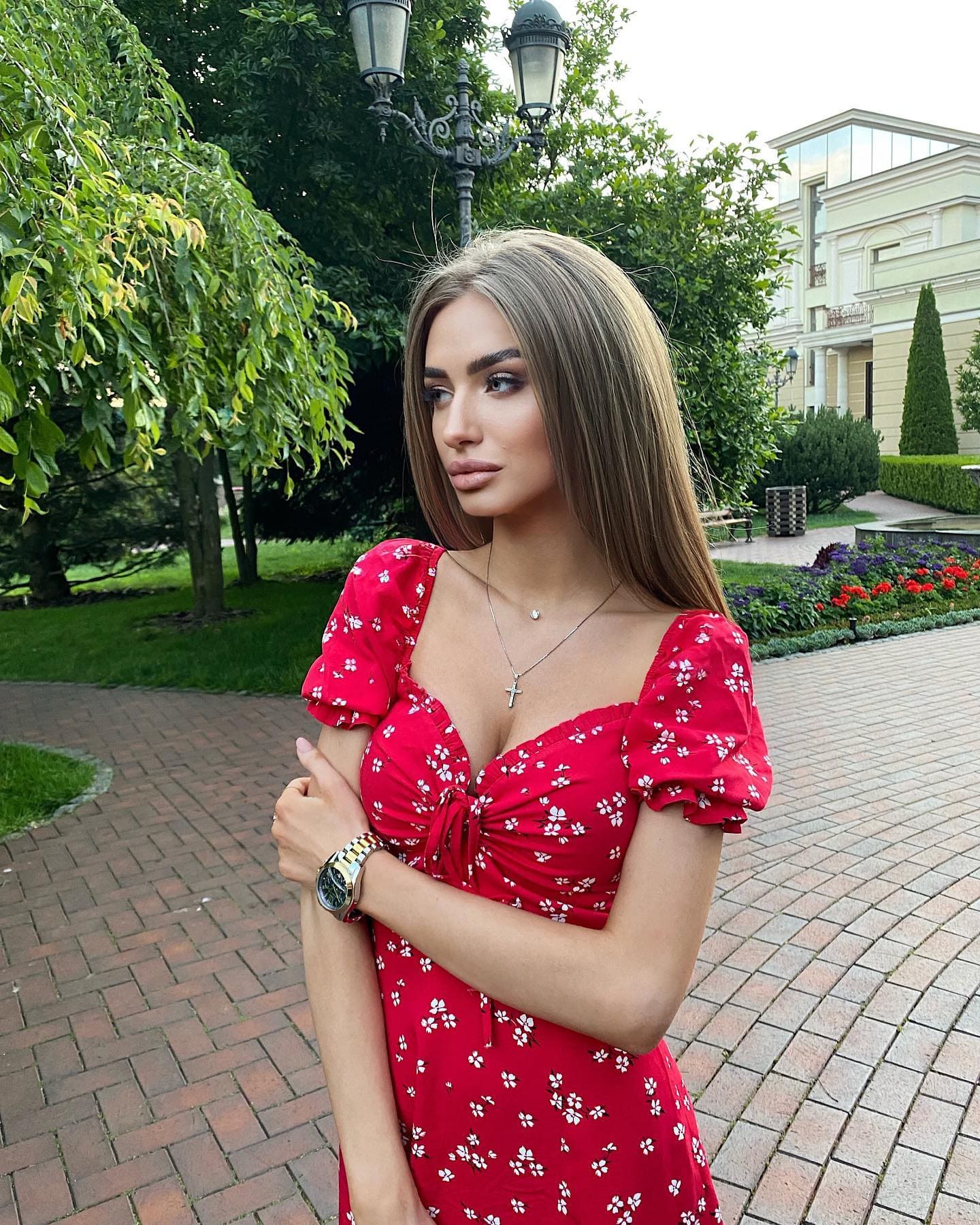 A woman with straight brown hair stands on a brick walkway in a red floral dress, arms crossed, looking to the side.