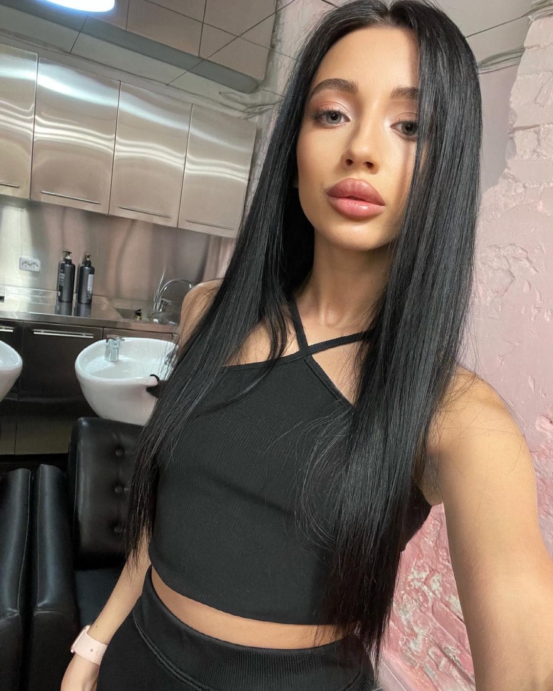 A woman in a black strappy crop top and high-waisted bottoms poses in a salon with white basins and metal cabinets.