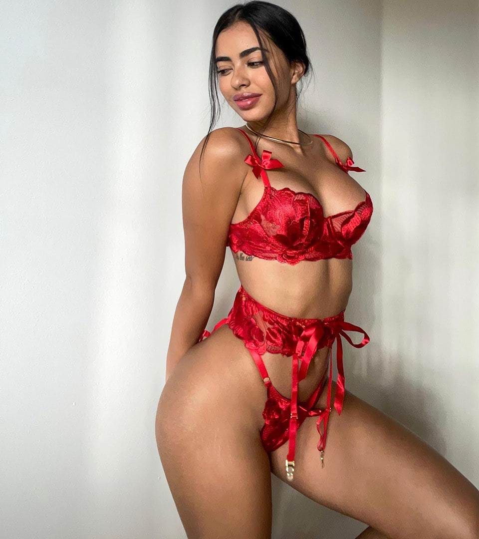 A woman in a red lace lingerie set with ribbon ties and a thin choker stands in a three-quarter pose against a plain light wall.