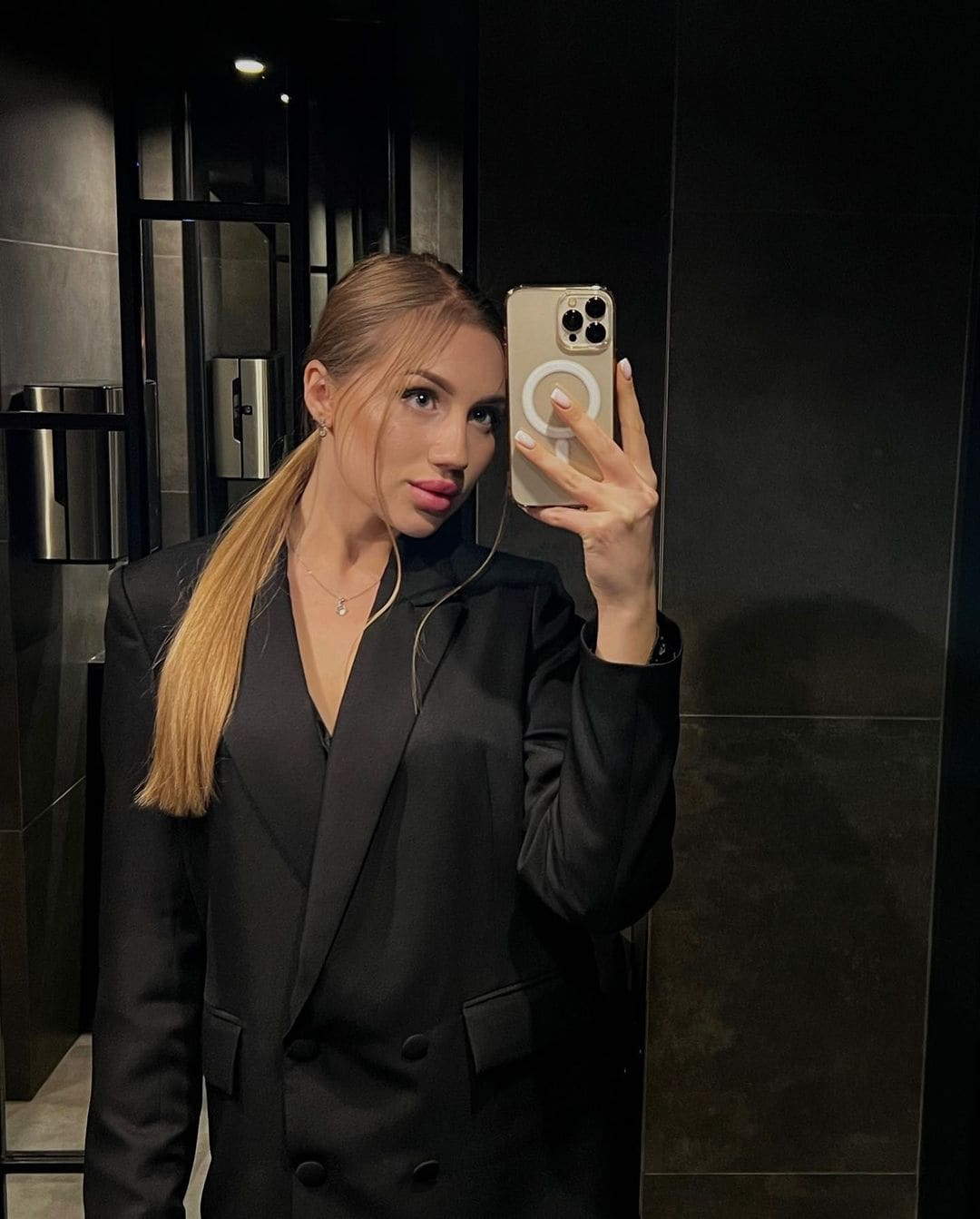 Mirror selfie in a black double-breasted blazer, hair in a side ponytail, in a dark tiled room.