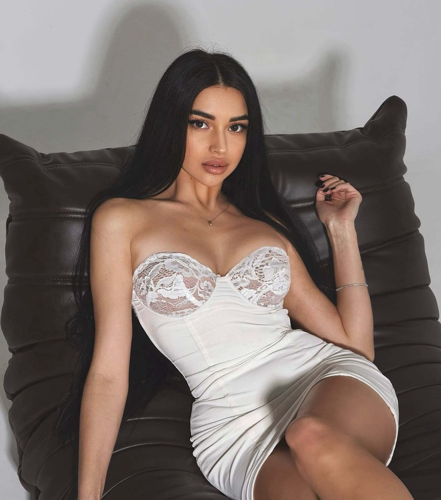 A woman reclines on a dark leather chair wearing a short white strapless dress with lace detailing, one hand raised near her shoulder.