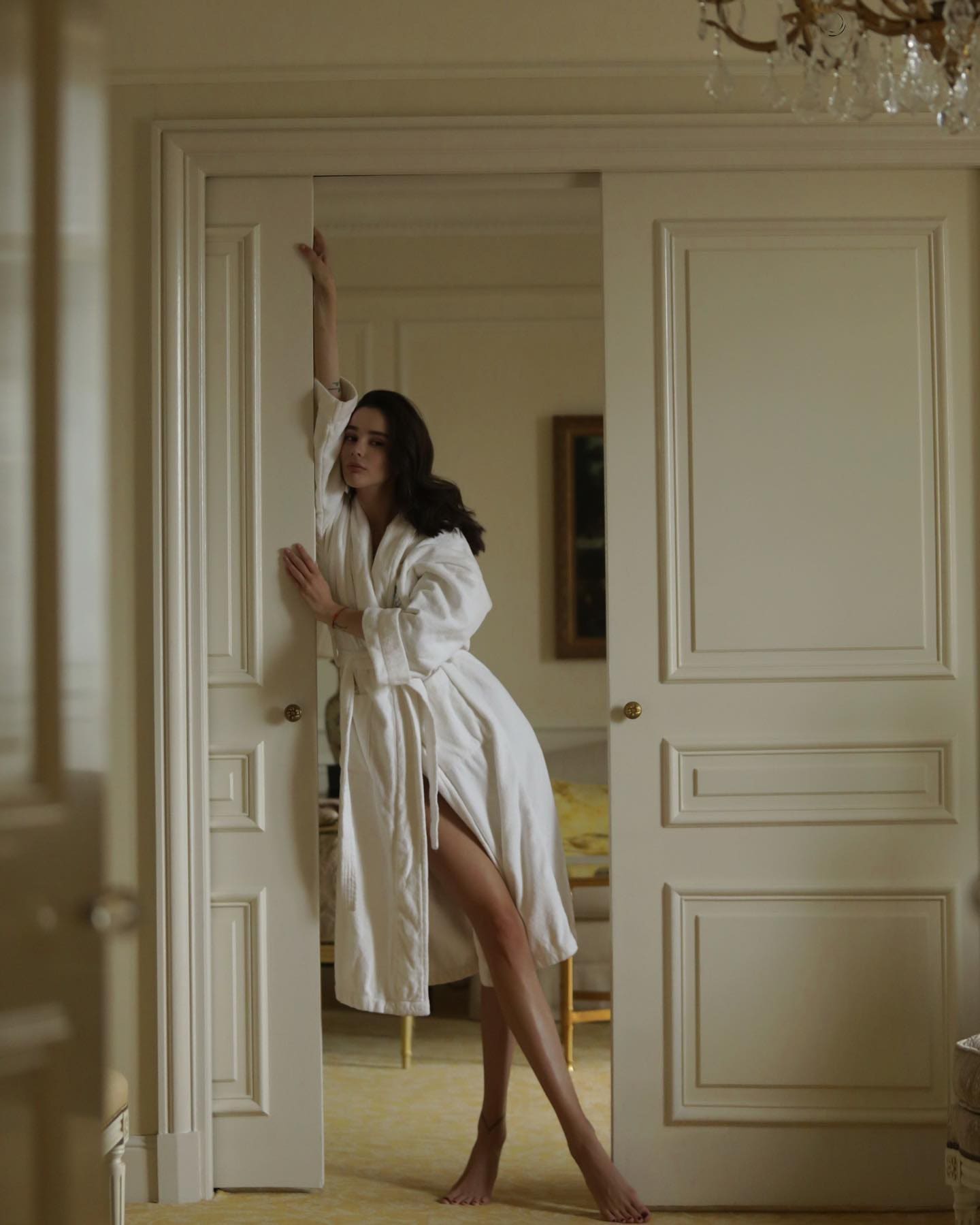 A woman stands barefoot in a doorway wearing an open white bathrobe, holding the frame with one arm and the door edge with the other.