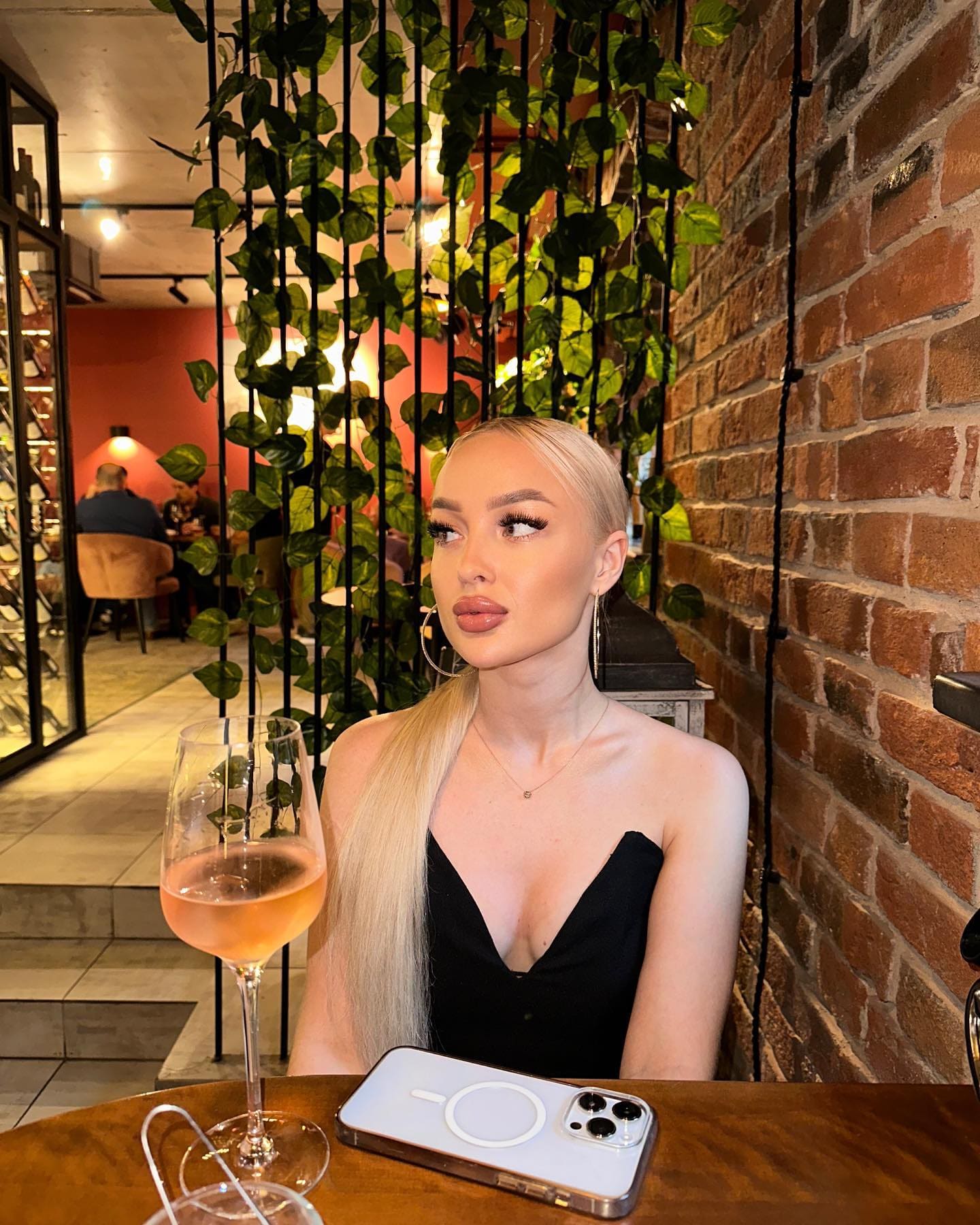 Seated against a brick wall with vines, she wears a black strapless top, with rosé wine and a smartphone on the wooden table.