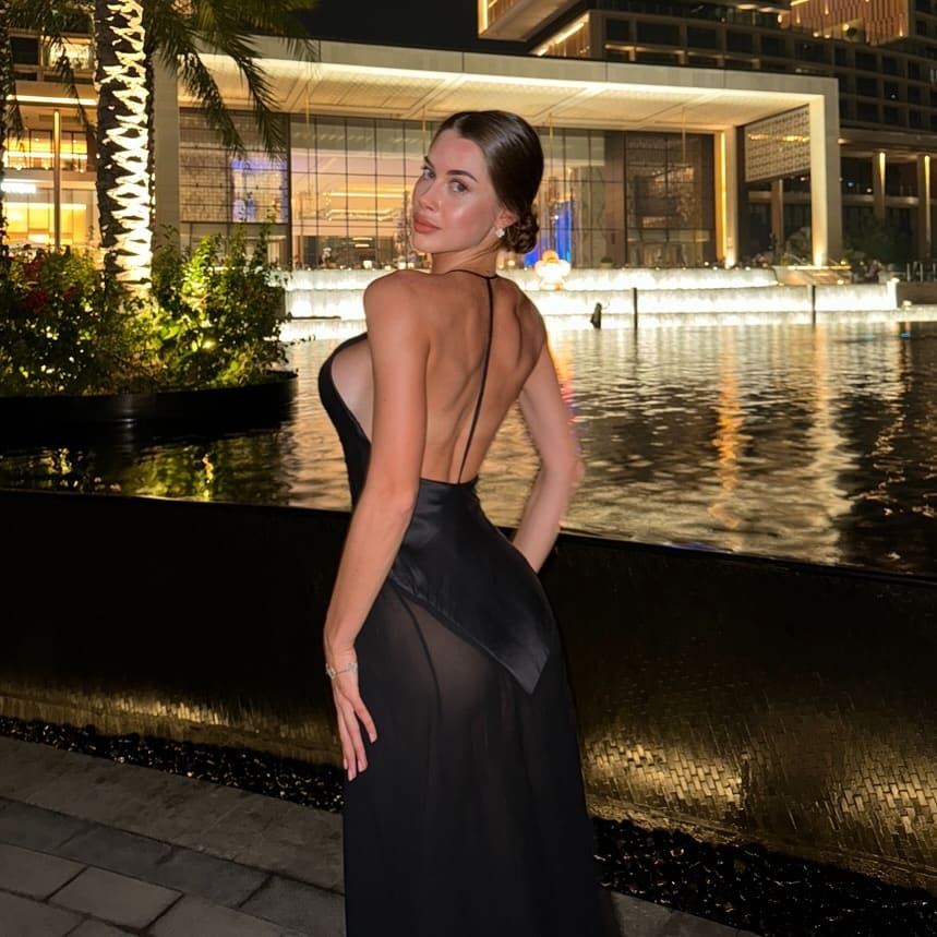 Profile view of a woman in a black evening gown with an open back, standing beside a lit water feature at night.