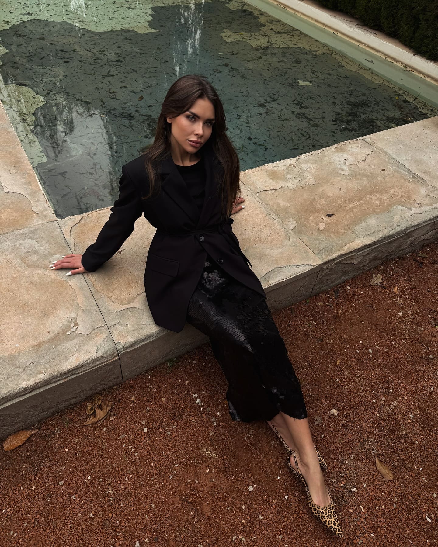 A woman sits on a stone ledge by water, wearing a black blazer, textured midi skirt, and leopard-print flats, hands resting on her lap.