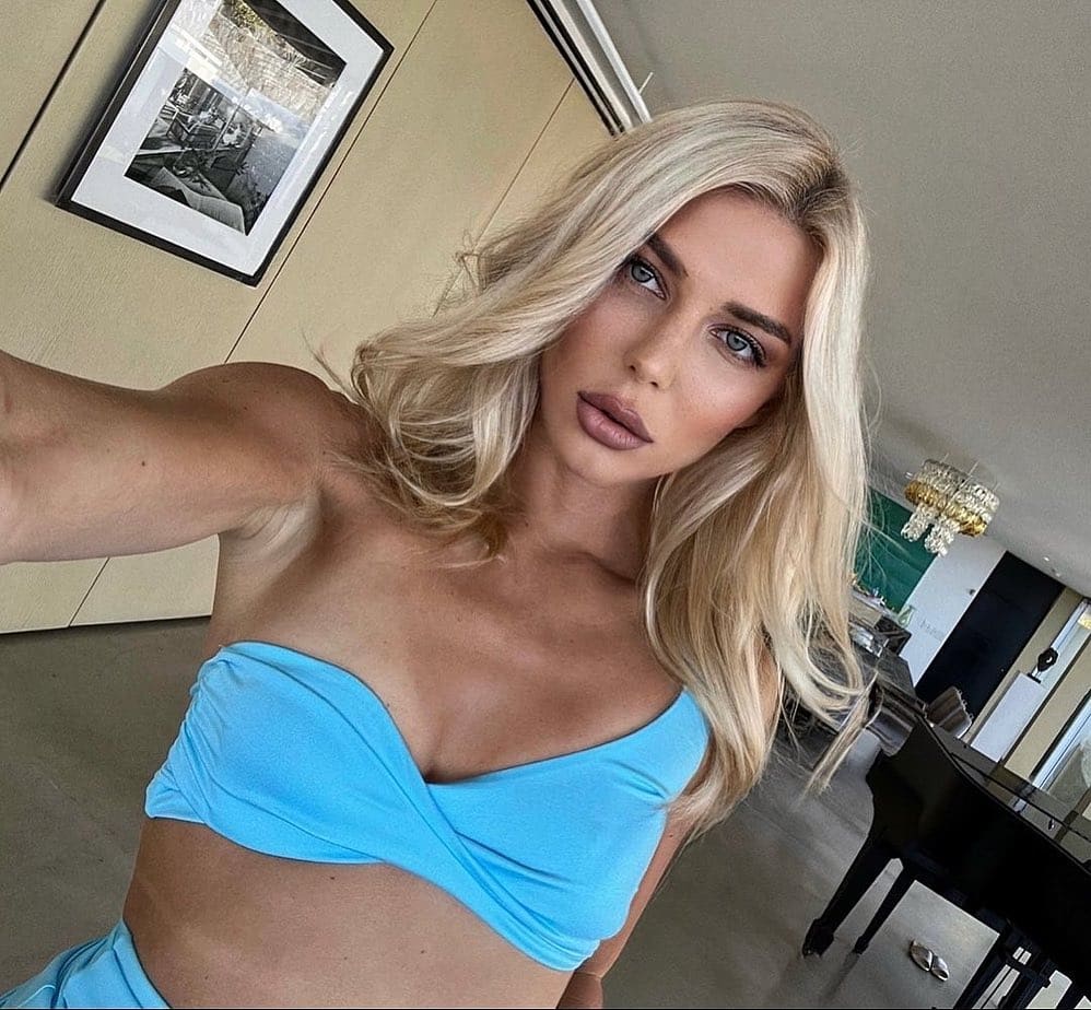 A selfie of a blonde woman in a light blue strapless top, with a framed black-and-white photograph and a grand piano visible in the indoor background.