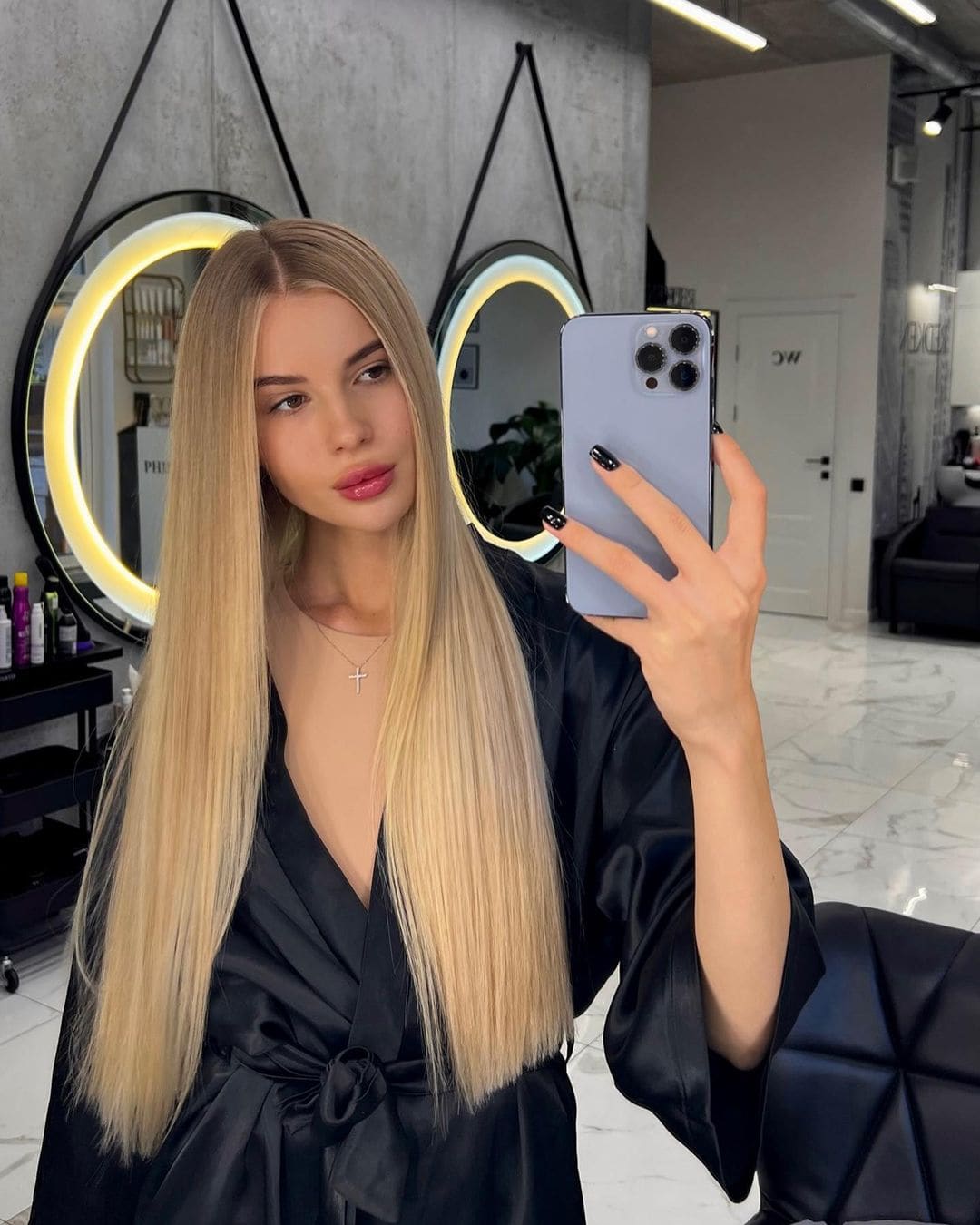 A mirror selfie in a salon shows a woman in a black satin robe with a deep V-neck and cross necklace, holding a light blue phone.