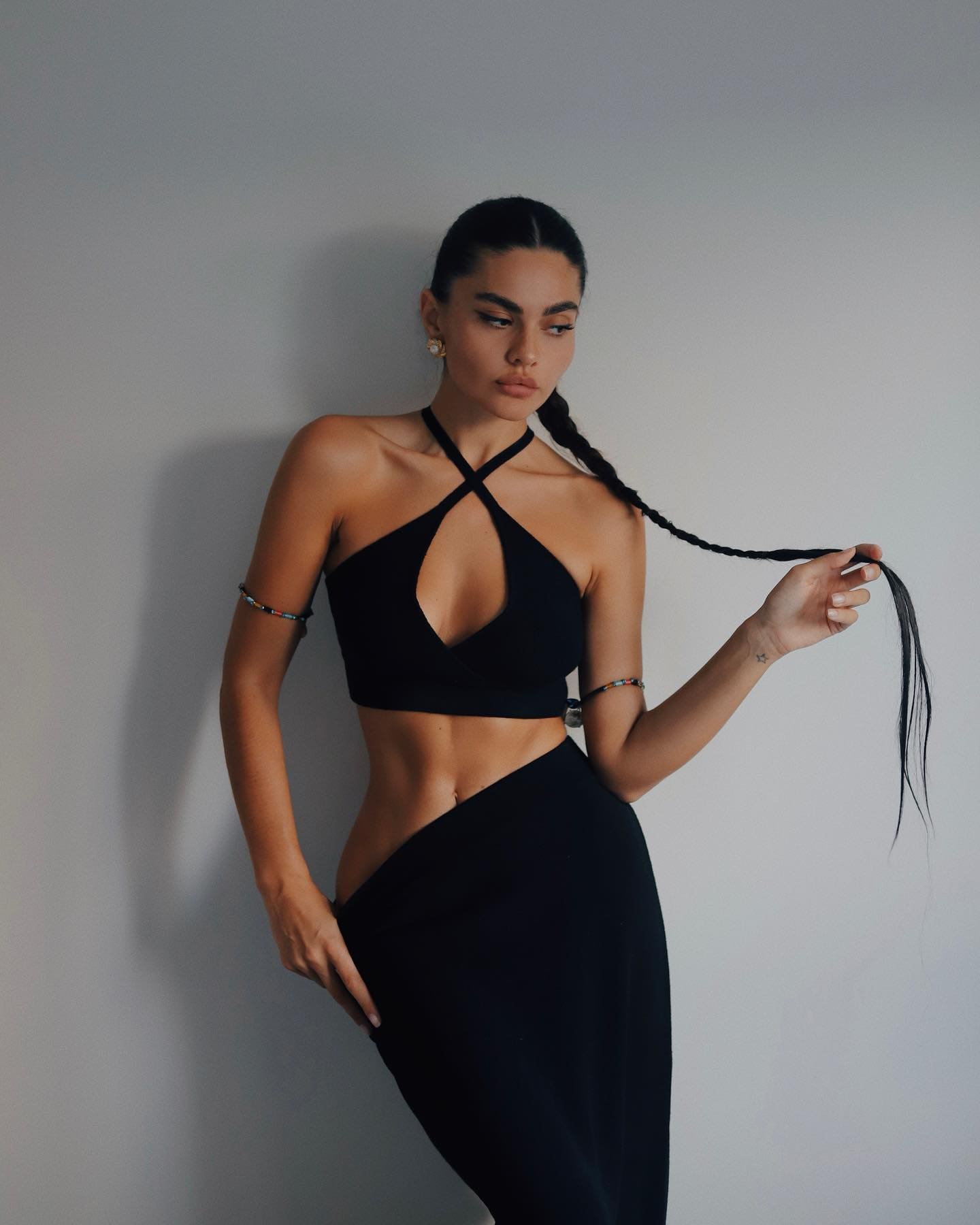 Mia stands against a gray wall in a black halter crop top and high-waisted skirt, holding her long dark braid.