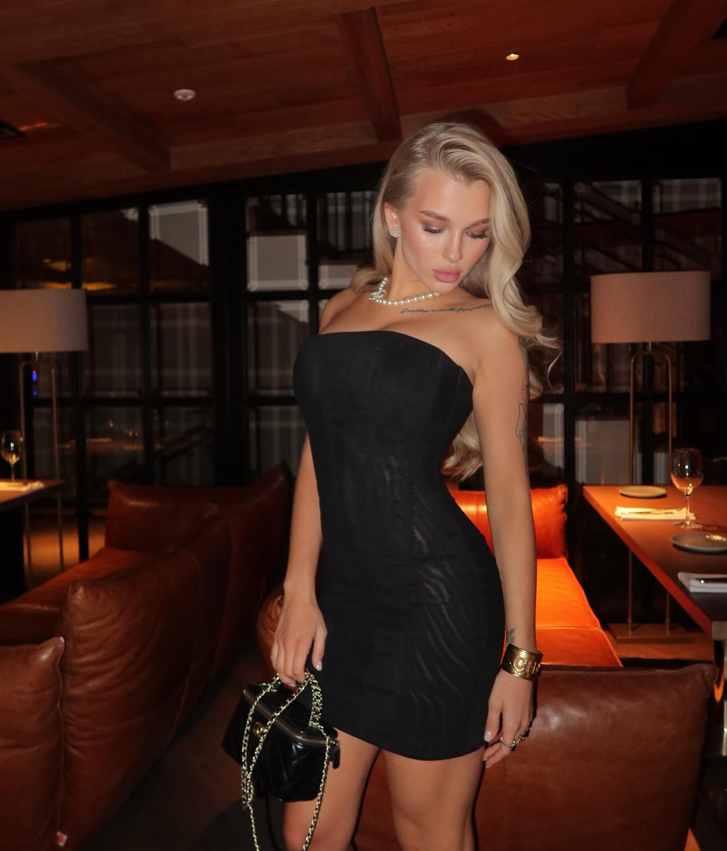 A blonde woman stands in a dimly lit restaurant wearing a black strapless mini dress, pearl necklace, and gold cuff, holding a small black handbag.