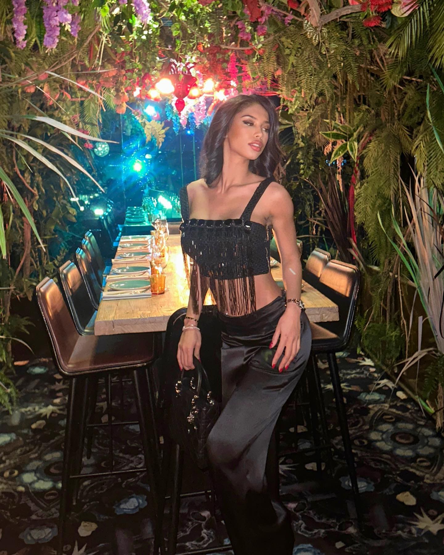 A woman sits on a high stool in a plant-filled venue, wearing a black fringed crop top and satin trousers while holding a black handbag.