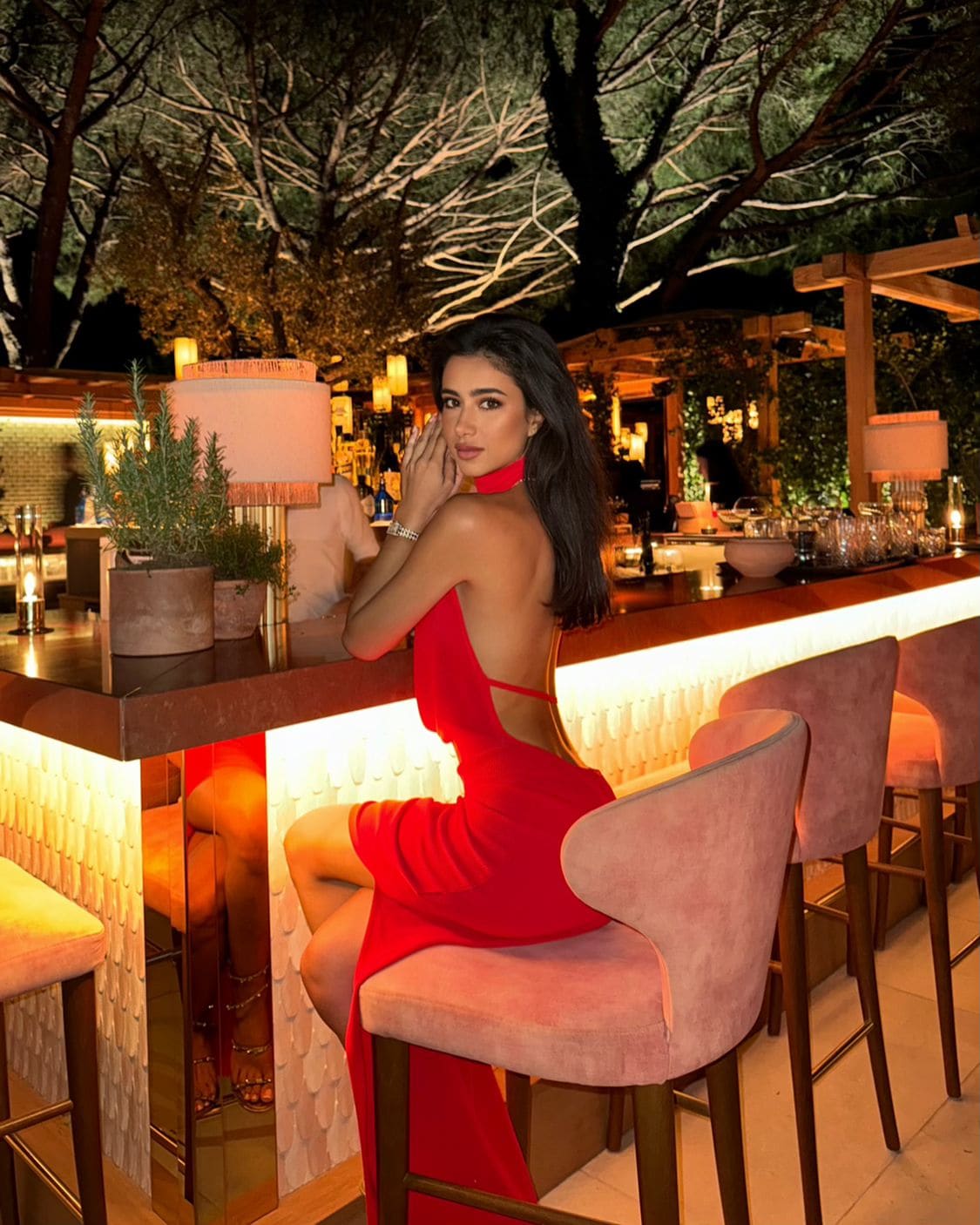 A woman sits on a pink stool at an outdoor bar at night, wearing a red backless dress with a slit, hand near her face.