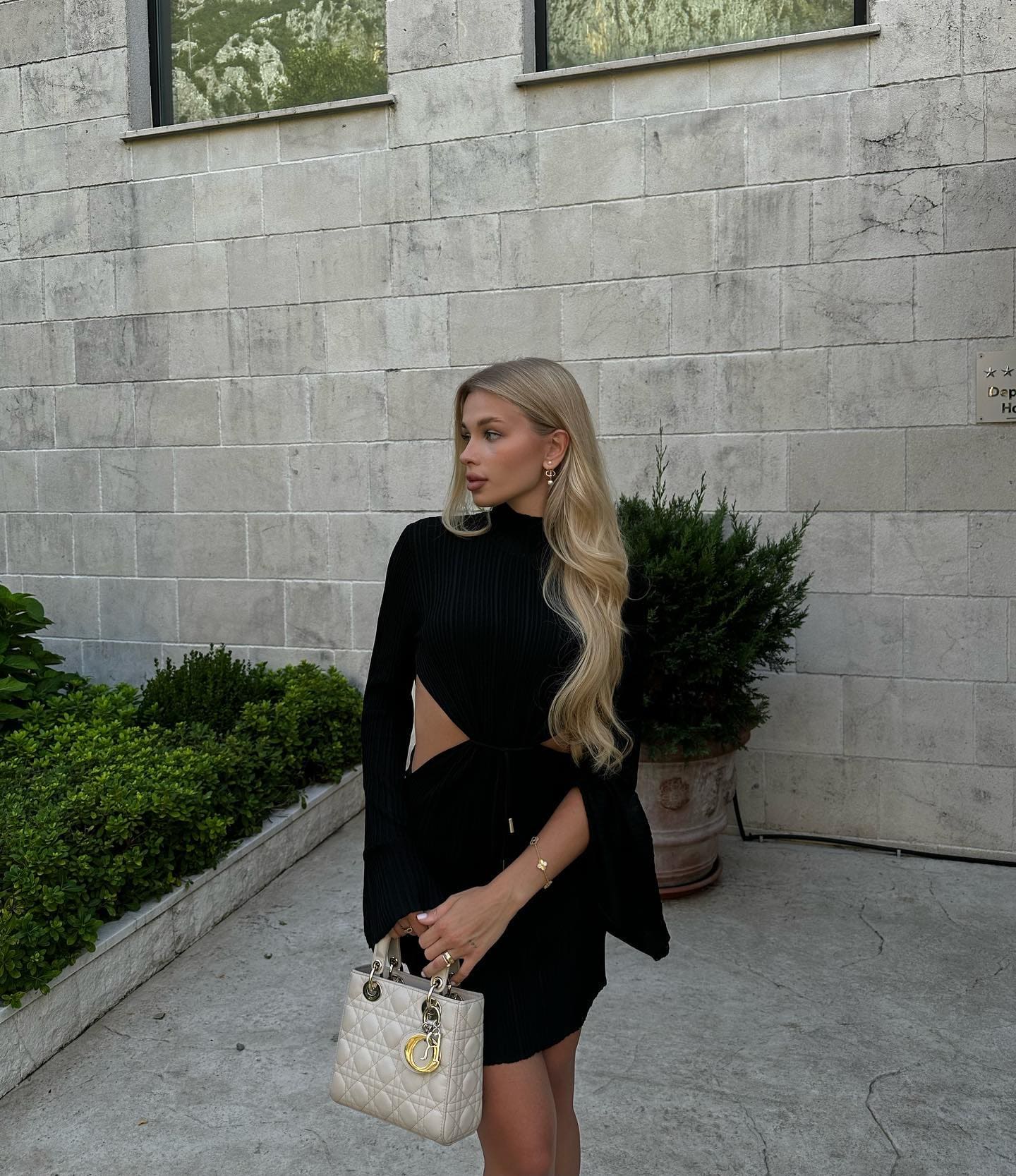 Profile view of a blonde woman in a black ribbed top with a side cutout and short skirt, holding a white quilted handbag against a grey stone wall.