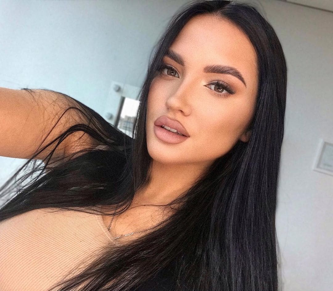 Close-up selfie of a woman with long dark hair in a sheer nude top, posing indoors against a plain light-colored wall.
