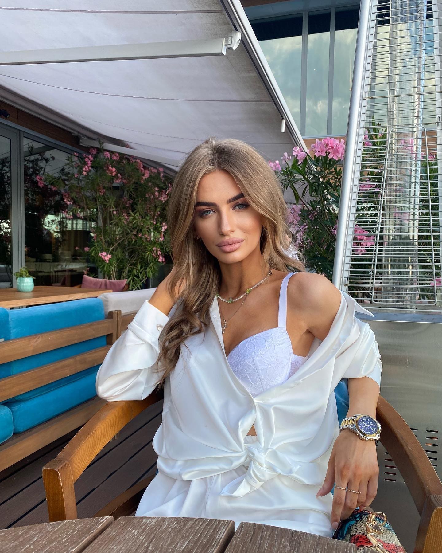 Love sits at an outdoor wooden table wearing a white satin shirt over a lace bralette, with pink flowers and a building behind her.