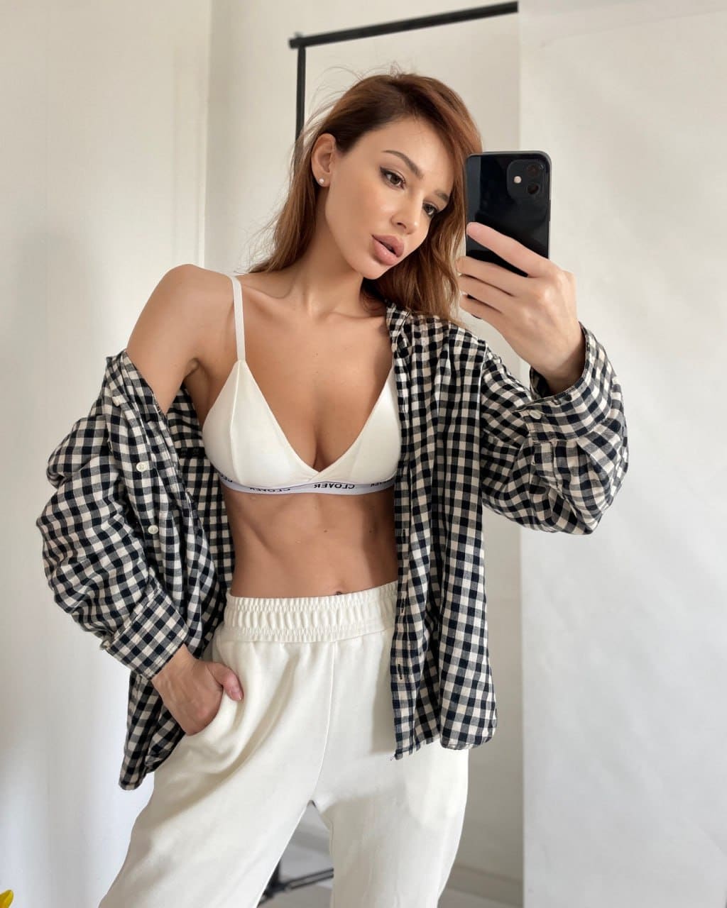 Woman in white bra, sweatpants, and open checkered shirt takes a mirror selfie in a white room.