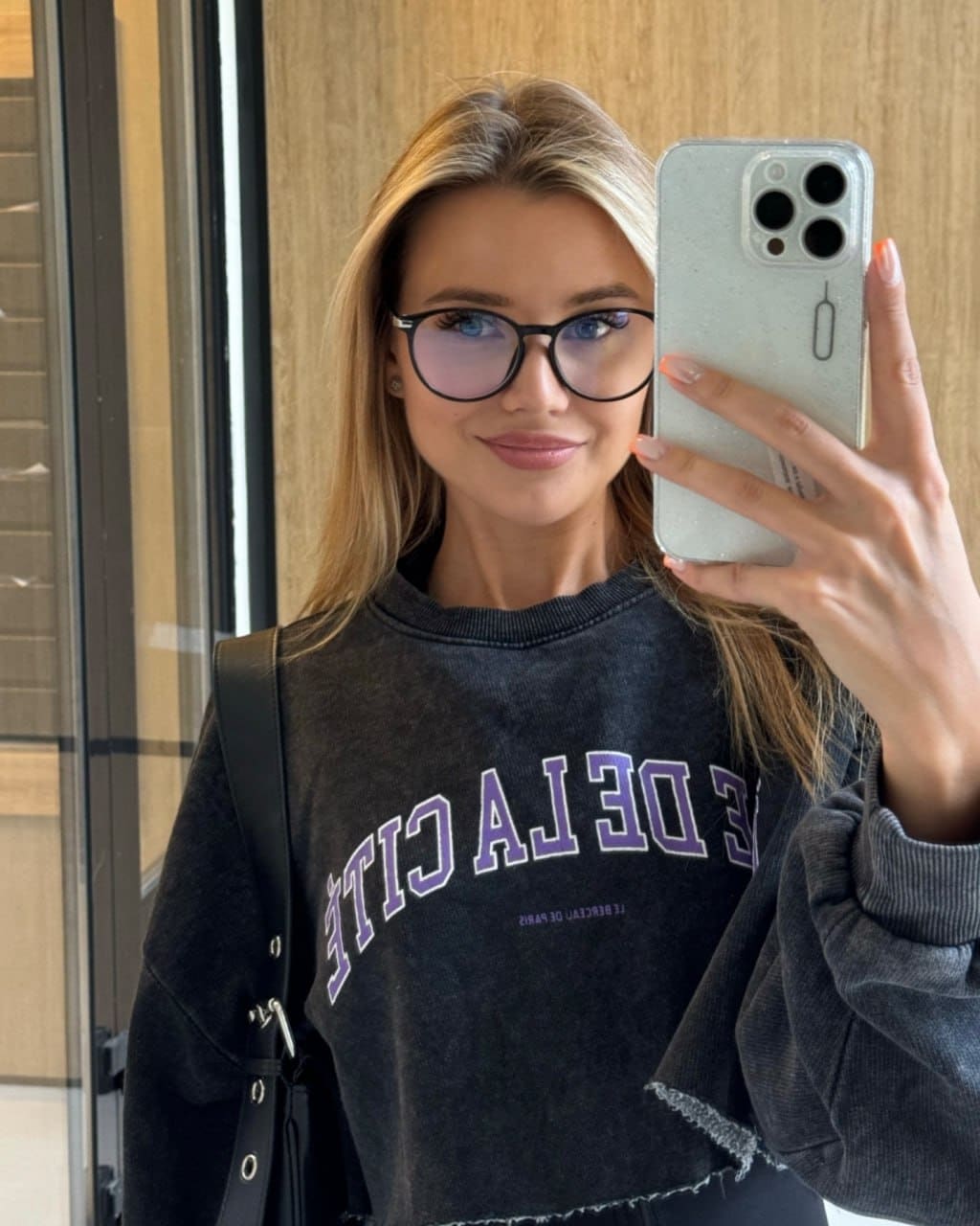Susanna wears glasses, a grey sweatshirt and black bag, taking a mirror selfie by a wooden wall and glass door.
