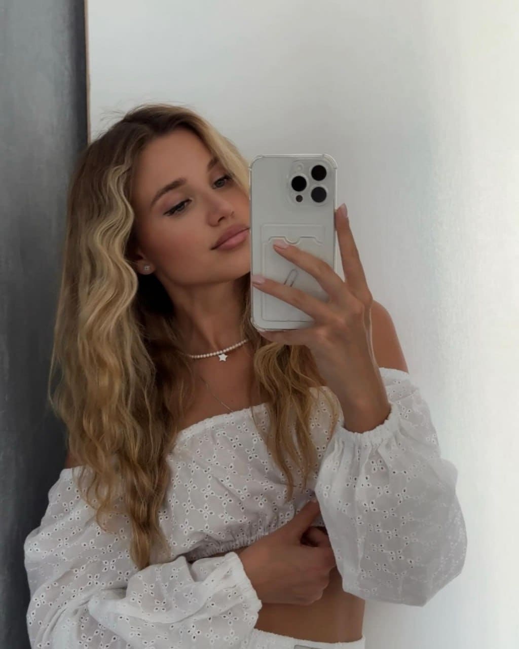A blonde woman in a white eyelet crop top and necklace holds a phone for a mirror selfie against a plain wall.